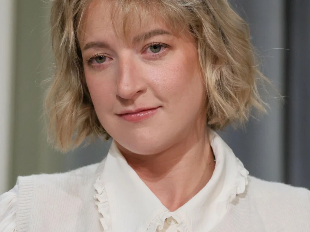 Blondynka o krótkich, falowanych włosach w białej koszuli z falbankami, patrząca z delikatnym uśmiechem na twarzy. Tło rozmyte, neutralne, skupiające uwagę na twarzy kobiety.