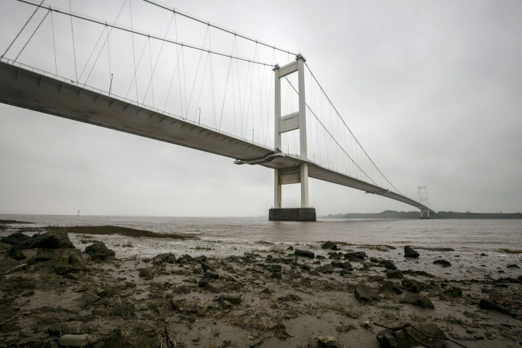 Duży most wiszący rozciągający się nad szeroką rzeką przy pochmurnym, szarym niebie. Widać masywne filary oraz liny podtrzymujące konstrukcję, brzeg rzeki jest kamienisty i błotnisty.