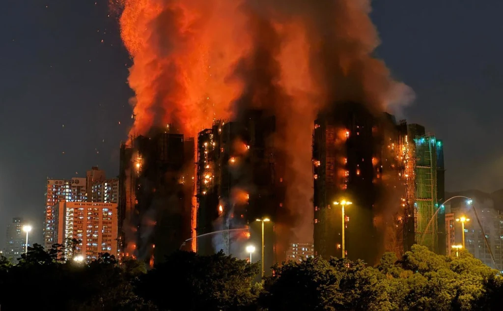 Wysoki budynek objęty jest silnym pożarem, z dachu i okien wydobywają się duże płomienie i kłęby gęstego, ciemnego dymu. W tle widoczne są inne miejskie wieżowce oświetlone nocą, a pod budynkiem znajdują się drzewa i latarnie uliczne.