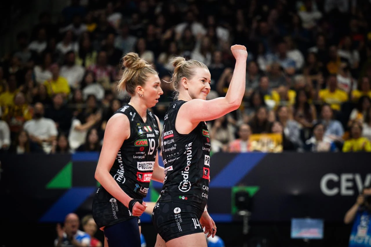 Joanna Wołosz w barwach Imoco Conegliano Dwie zawodniczki siatkarskie w czarnych strojach zespołu obchodzą ważny moment podczas meczu, jedna z nich unosi pięść w geście zwycięstwa lub radości, w tle widoczna jest publiczność skupiona na trybunach.