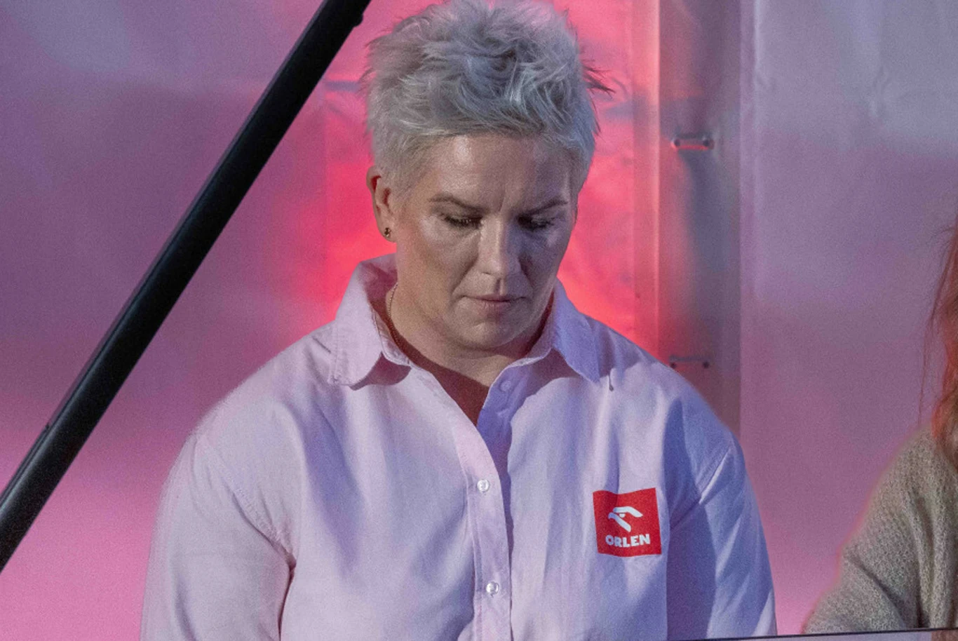Anita Włodarczyk Osoba o krótkich, siwych włosach w jasnej koszuli z naszywką Orlen, patrząca w dół, oświetlona różowym światłem stojącym na tle jasnej ściany.