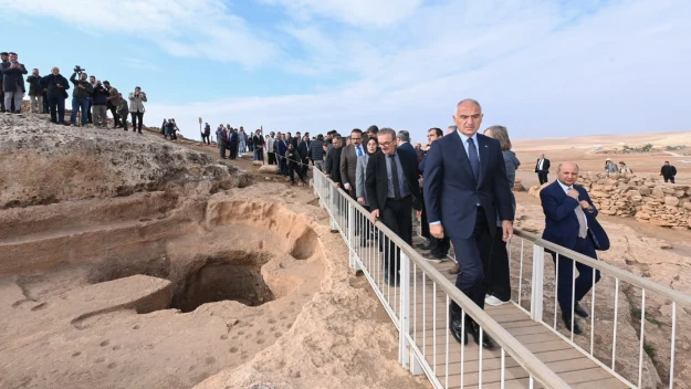 Grupa elegancko ubranych osób przechodzi po wąskiej kładce ponad wykopem archeologicznym, na tle rozległego, pustynnego krajobrazu i zebranej licznie publiczności na wzniesieniu.