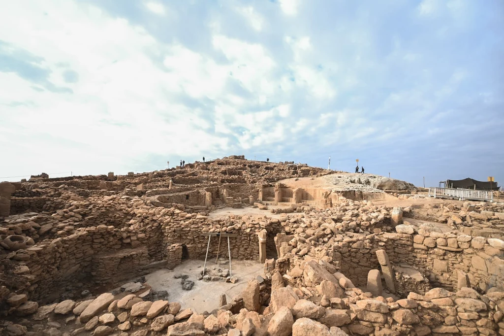 Projekt Taş Tepeler obejmuje 12 neolitycznych stanowisk w Turcji. Badacze przedstawili nowe znaczące odkrycia. Ruiny starożytnego stanowiska archeologicznego z kamiennymi murami i fundamentami budynków na tle jasnego nieba. Na szczycie wzniesienia widoczne osoby zwiedzające teren wykopalisk.
