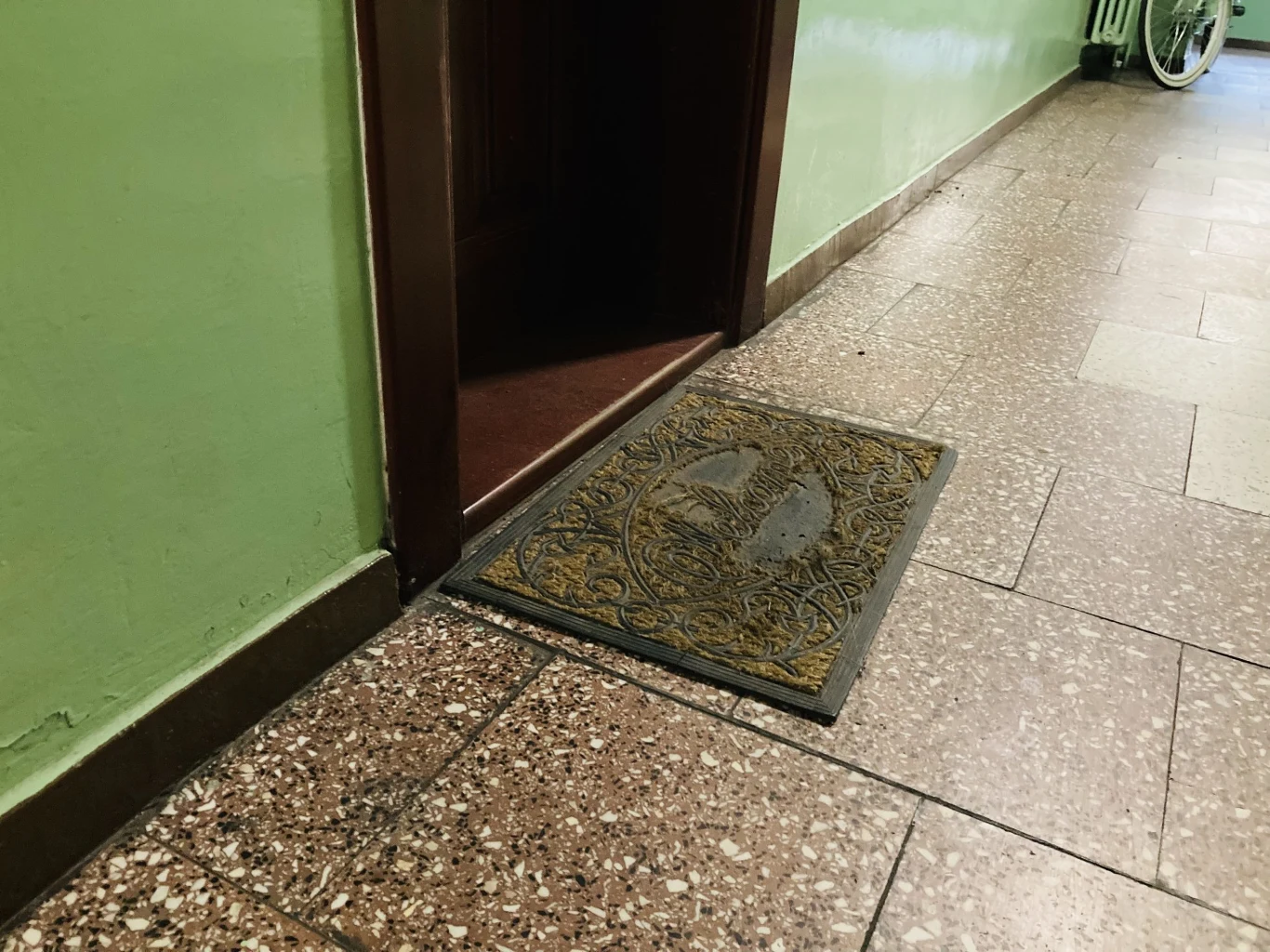 Ta niepozorna rzecz może mieć niebagatelny wpływ na twoje finanse Stara wycieraczka z wyraźnymi śladami zużycia leży przy otwartych ciemnych drzwiach w korytarzu wyłożonym brązowymi płytkami, ściany pomalowane na jasnozielono.