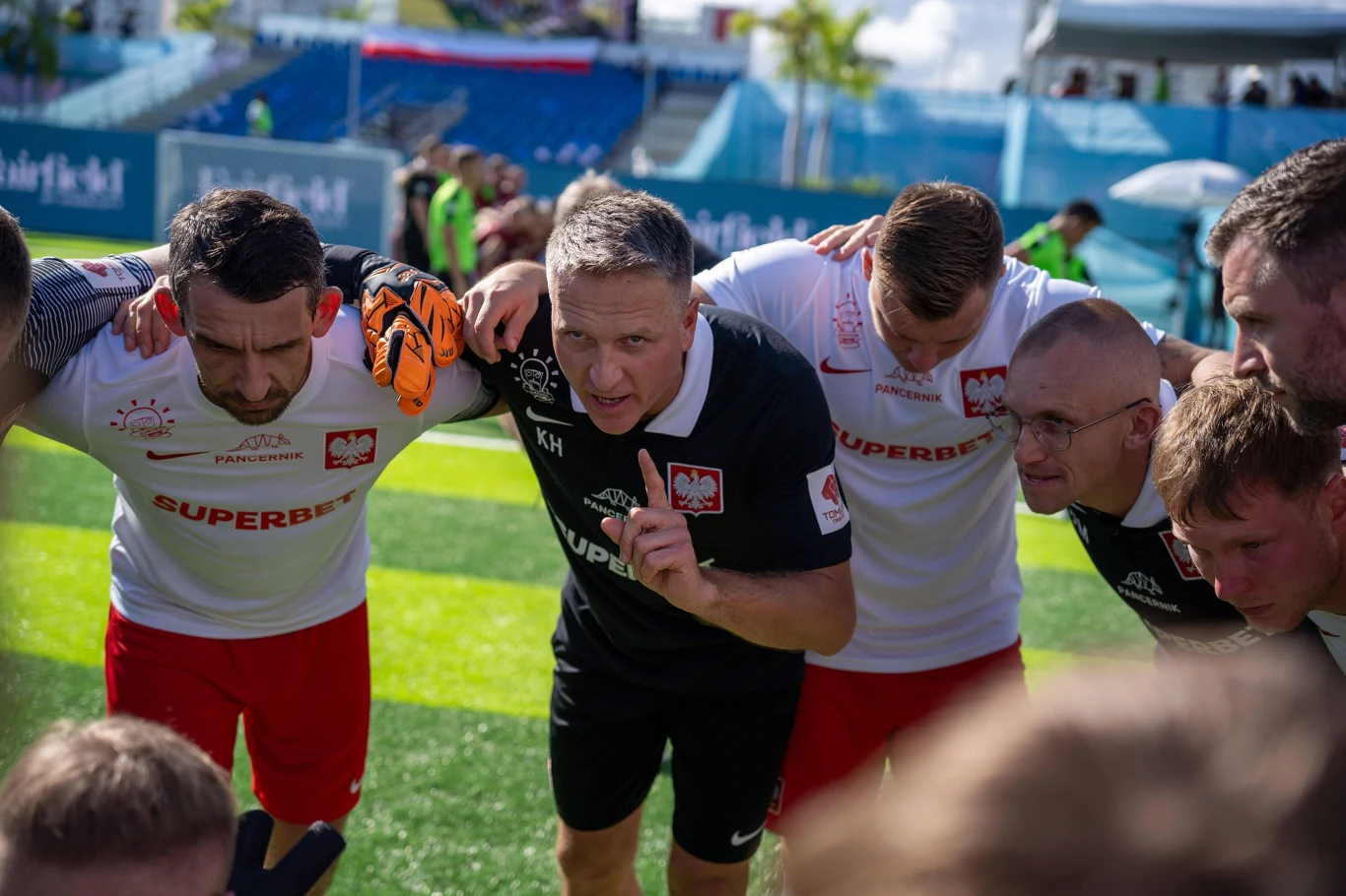 Klaudiusz Hirsch poprowadził Polaków do złotego medalu MŚ Grupa polskich piłkarzy wraz z trenerem zbierają się w kręgu na boisku, intensywnie skupieni i słuchający instrukcji, z widocznymi sportowymi strojami i emblematami narodowymi.