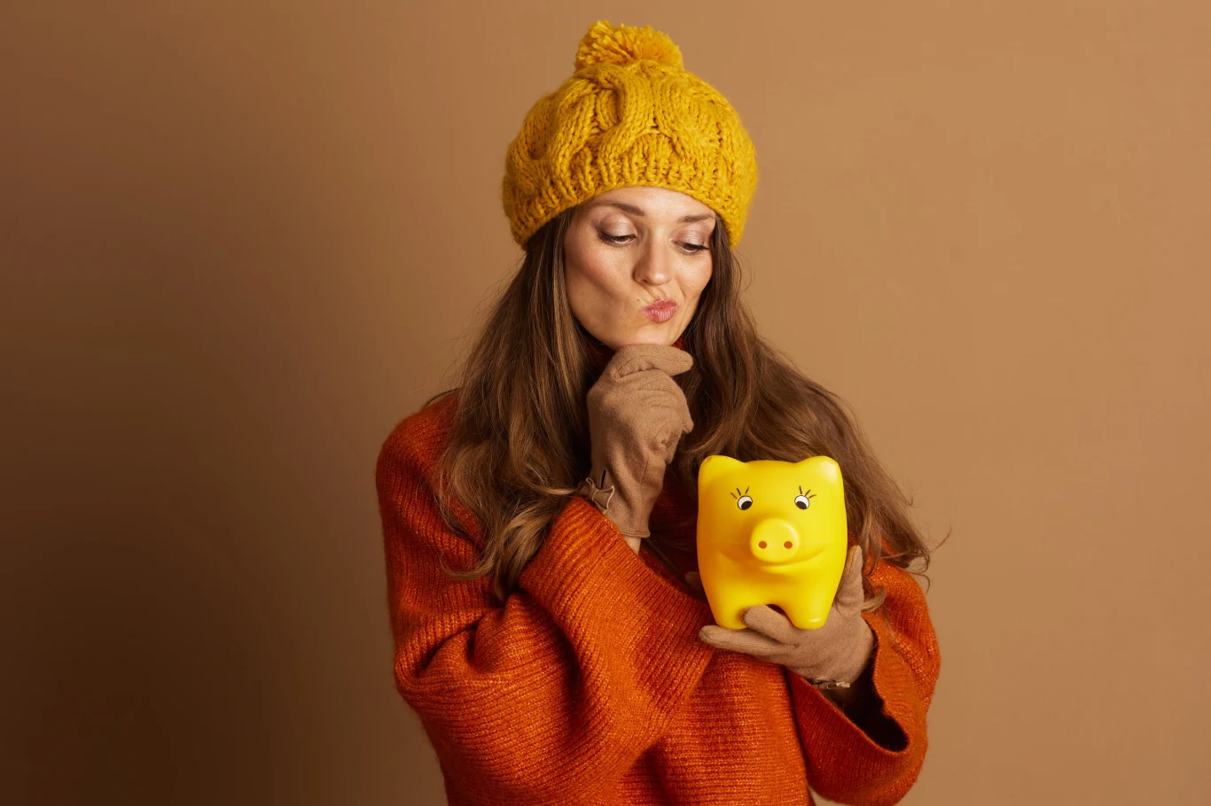 Numerologiczna analiza grudniowa: co czeka twoją karierę i finanse Kobieta w żółtej czapce i pomarańczowym swetrze trzyma żółtą skarbonkę w kształcie świnki i patrzy na nią zamyślona.