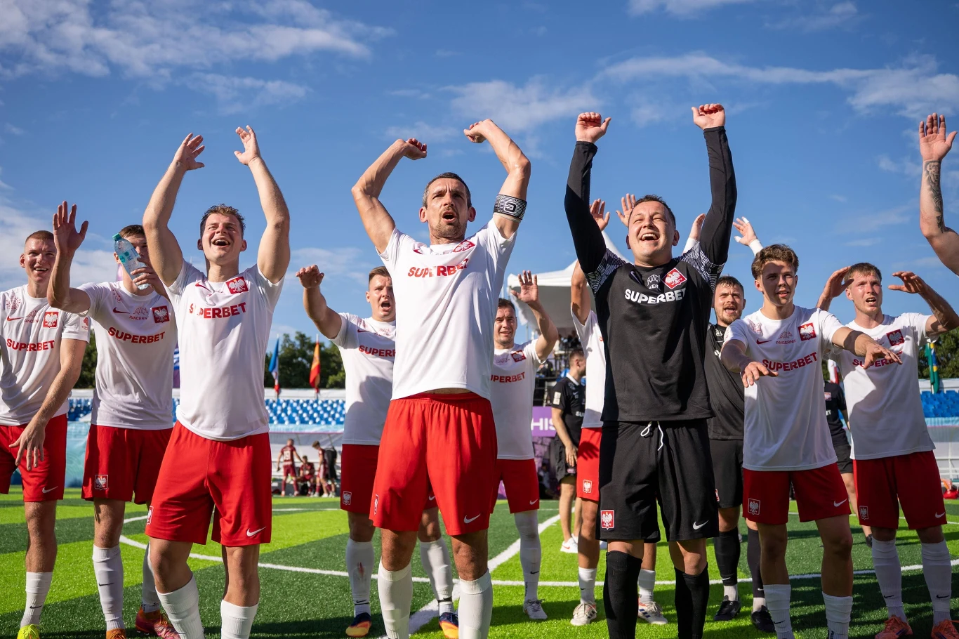 Polska została MŚ w socca Grupa piłkarzy w jednakowych strojach biało-czerwonych świętuje triumf na murawie boiska, unosi ręce w geście zwycięstwa, uśmiechnięci zawodnicy stoją razem na tle słonecznego nieba.