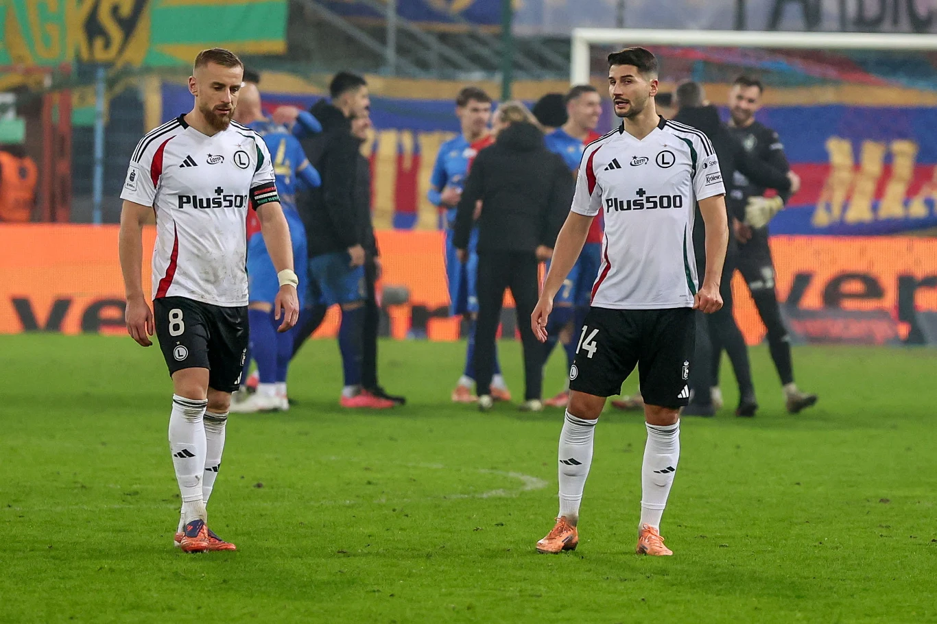 Piłkarze Legii Warszawa Dwóch piłkarzy w białych strojach Legii Warszawa spaceruje po murawie stadionu. W tle widoczna grupa zawodników w niebieskich strojach oraz banery i trybuny wypełnione kibicami.