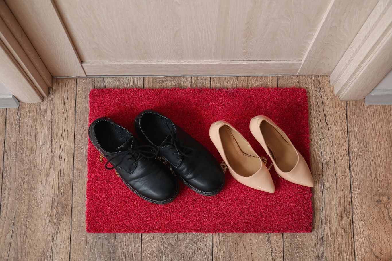 Niedopasowana do kierunku czerwona wycieraczka może porządnie "namieszać" w twoich finansach Para czarnych męskich butów oraz para beżowych damskich szpilek ustawione na czerwonym wycieraczce przy drzwiach na jasnej drewnianej podłodze.