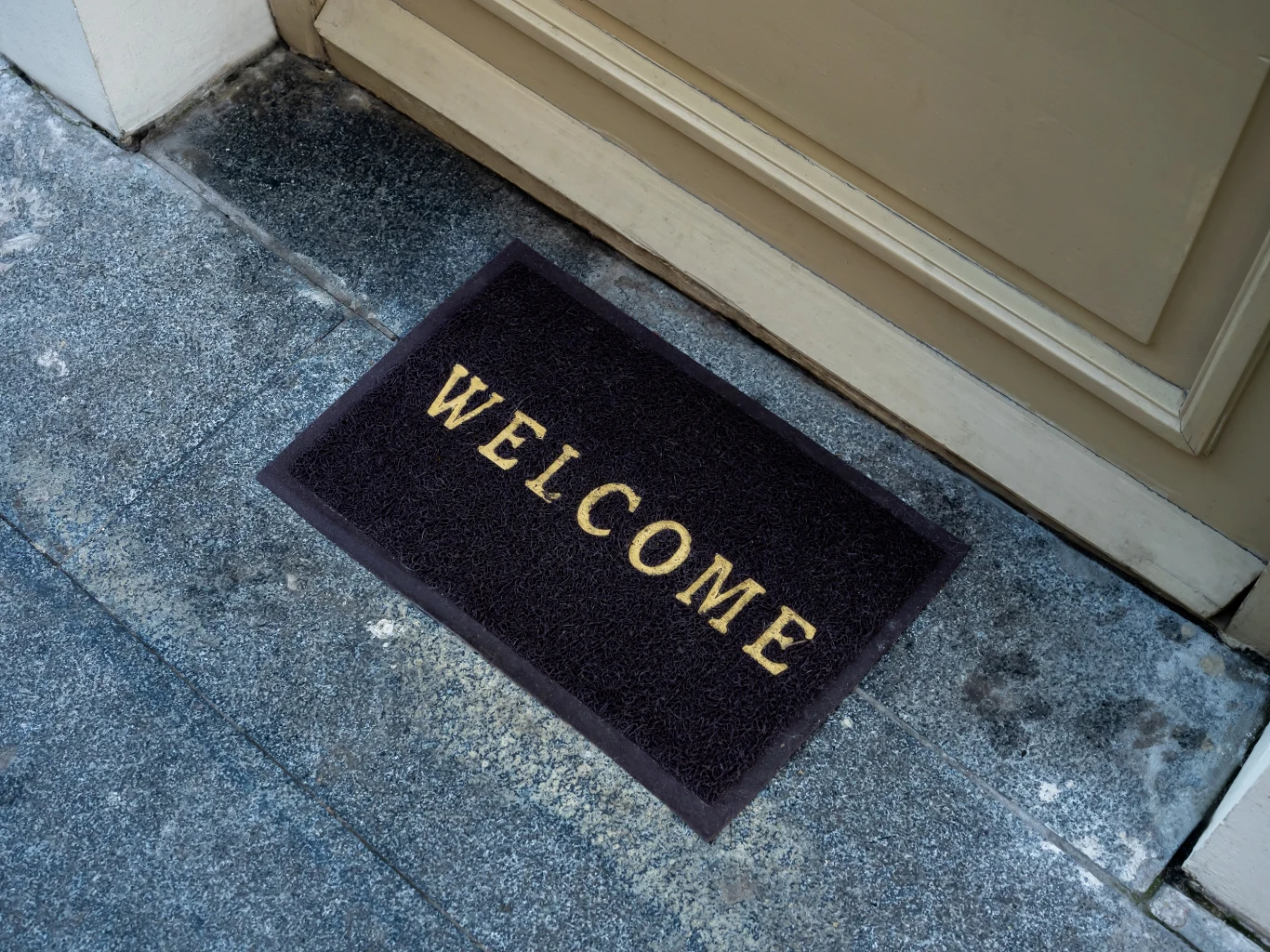 Czarne wycieraczki są bardzo popularne, ale nie dla każdego będzie to dobry wybór Czarna wycieraczka z napisem 'WELCOME' leży na szarej posadzce przy drzwiach wejściowych, powierzchnia wokół niej jest lekko zabrudzona.