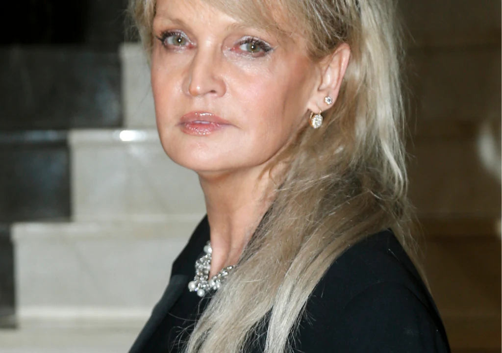 Adrianna Biedrzyńska Blondynka o długich włosach w eleganckiej czarnej kreacji, z biżuterią na szyi i w uszach, stoi na tle marmurowych schodów, spoglądając poważnie w stronę obiektywu.