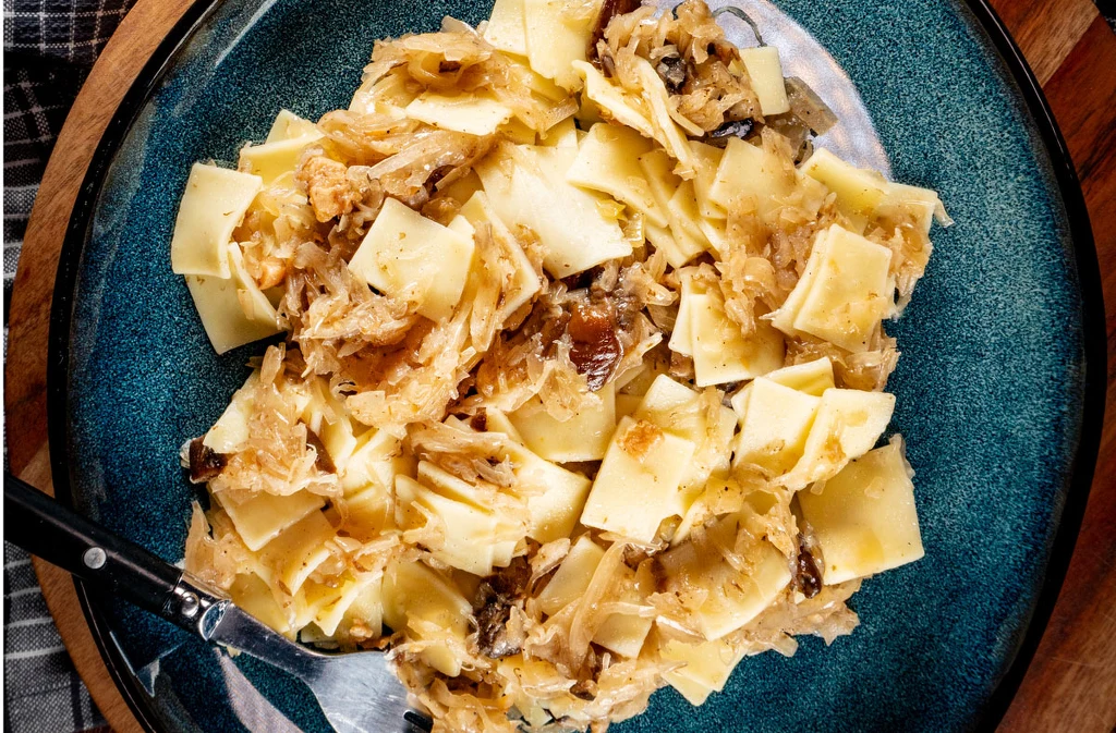 Makaron typu łazanki wymieszany z duszoną kapustą i grzybami, podany na niebieskim talerzu, sztućce przy talerzu.