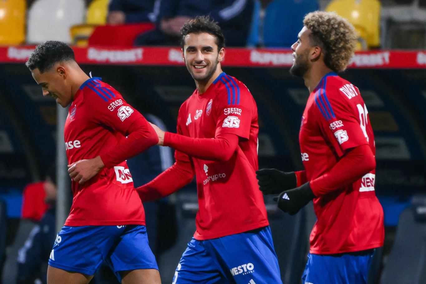 Ameyaw, Konstantopoulos i Diaby - Fadiga w barwach Rakowa Częstochowa Trzech piłkarzy w czerwonych koszulkach świętuje sukces na boisku piłkarskim, dwóch z nich ma krótkie włosy, jeden charakterystyczną bujną fryzurę, zawodnicy uśmiechają się i są wyraźnie zadowoleni.