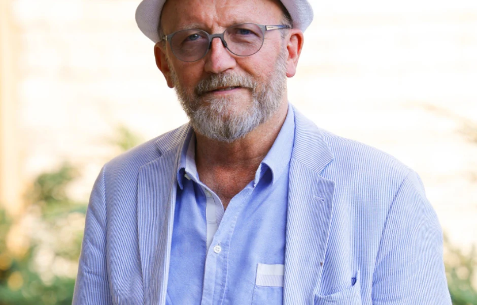 Artur Barciś Starszy mężczyzna z brodą i okularami w okrągłych oprawkach, ubrany w jasnoniebieską koszulę i marynarkę w drobny prążek, na głowie ma białą kapelusz, stoi na tle jasnego, rozmytego tła z delikatną roślinnością.