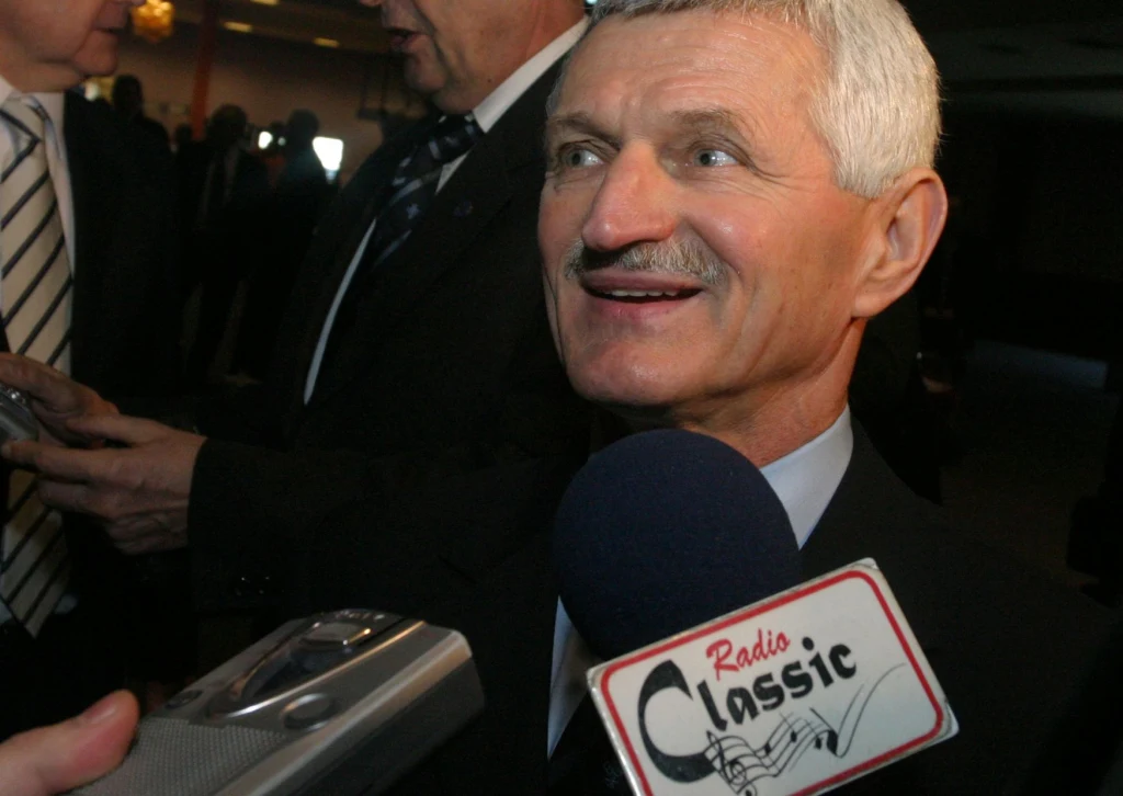 Zygmunt Smalcerz Mężczyzna w garniturze udzielający wywiadu, uśmiechnięty, przed nim trzymane są mikrofony oraz dyktafon, w tle widoczne inne osoby w eleganckich ubraniach.