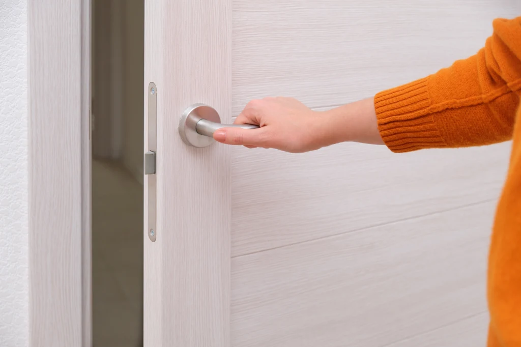 Ręka osoby w pomarańczowym swetrze chwyta za metalową klamkę białych, nowoczesnych drzwi.