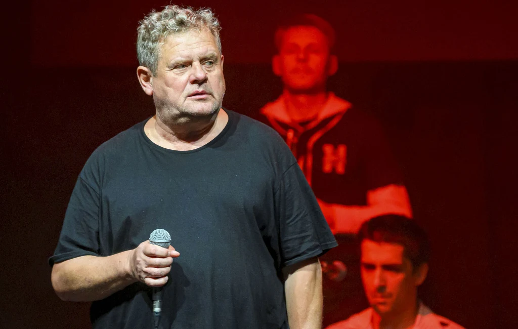 Kazik Staszewski opublikował nowy wpis ze szpitala Mężczyzna w średnim wieku w czarnej koszulce trzyma mikrofon, stojąc na scenie. Za nim dwie osoby w rozmytym tle, jedna ubrana w ciemną bluzę z literą H, czerwona scenografia podkreśla teatralny lub koncertowy charakter wydarzenia.