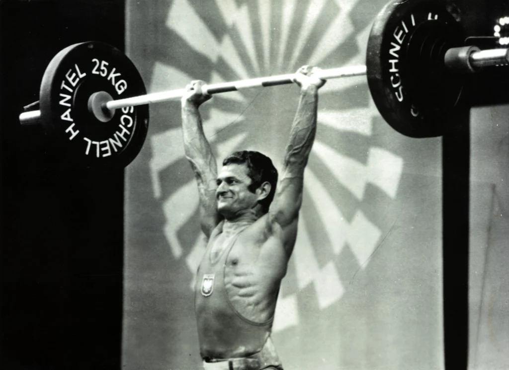 Zygmunt Smalcerz Zawodnik z odsłoniętym torsem podnosi sztangę z ciężarami nad głową, na tle graficznego motywu w kształcie promieni oraz herbu na piersi.