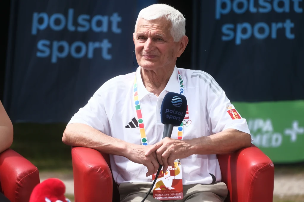 Zygmunt Smalcerz Starszy mężczyzna ubrany w biały sportowy strój i akredytację na szyi siedzi na czerwonym fotelu, trzyma mikrofon Polsat Sport, w tle widoczne banery tej stacji.