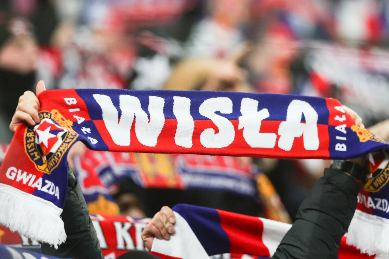 Szal klubowy Wisła Kraków trzymany w górze przez kibica na tle tłumu innych kibiców, dominują kolory czerwony, biały i niebieski.