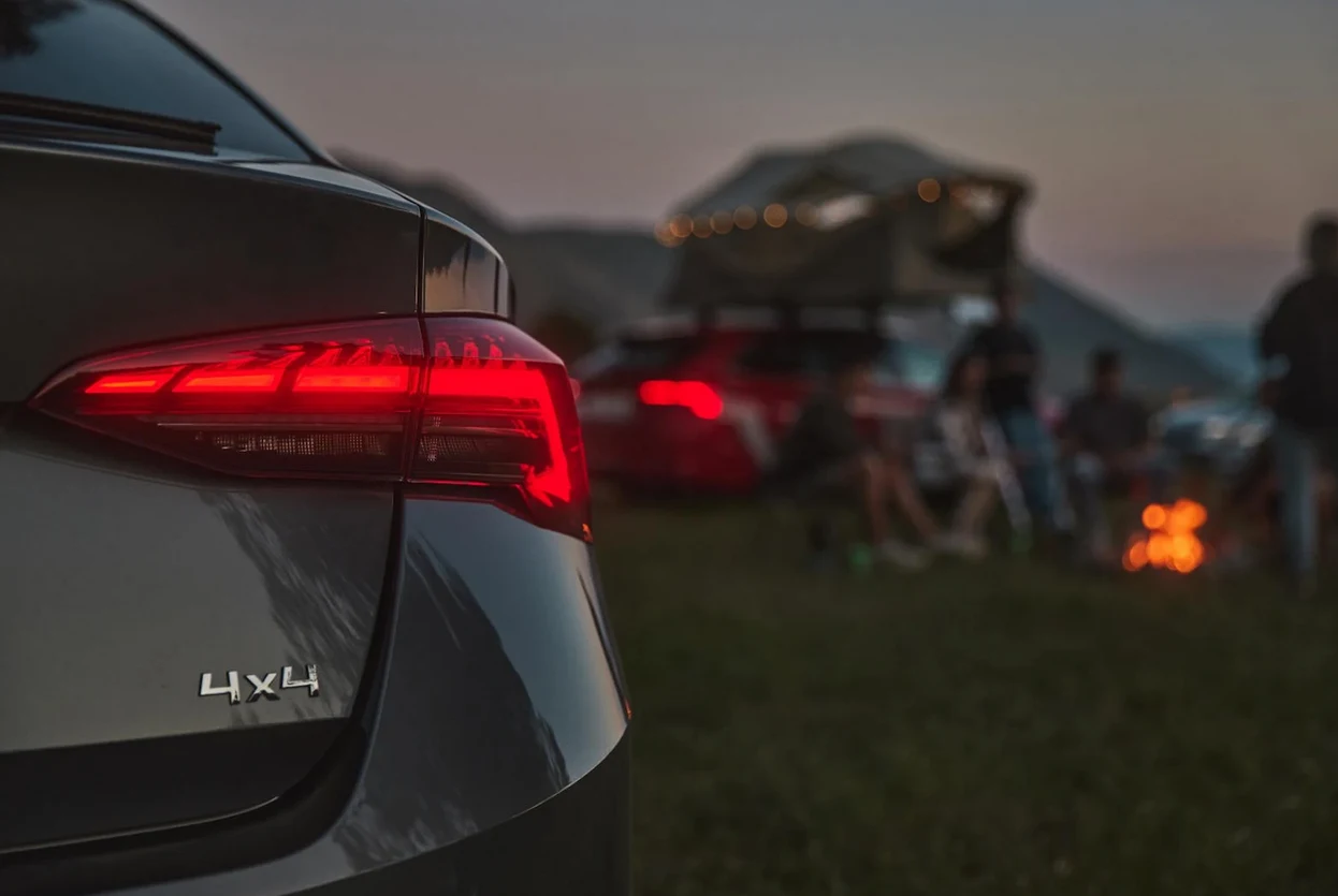 Odpowiedz na kilka pytań i przekonaj się, ile wiesz o napędzie 4x4 Tylna część czarnego samochodu z napędem 4x4, przy której rozmyta grupa ludzi siedzi obok ogniska na tle samochodu z namiotem dachowym w plenerze górskim o zmierzchu.