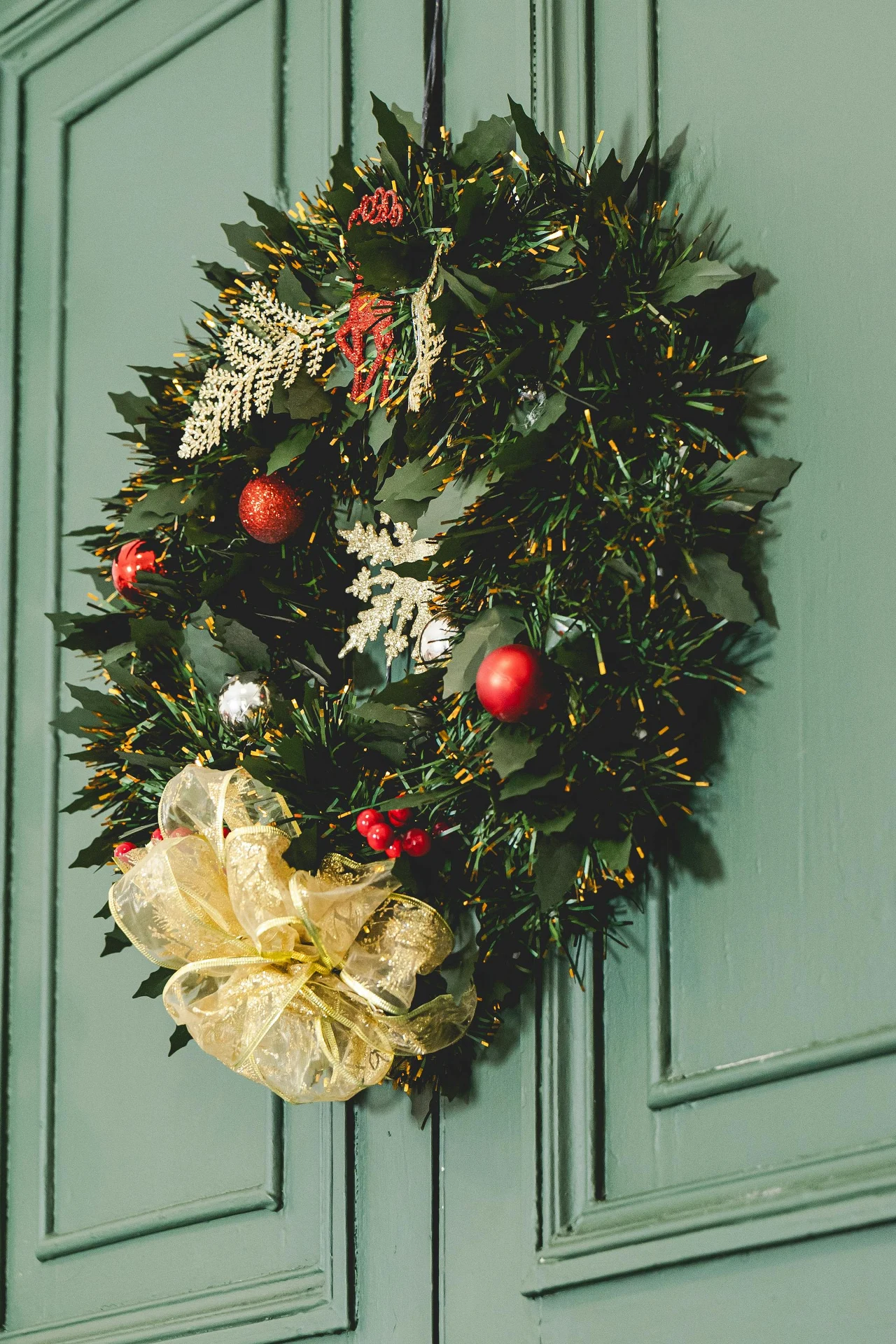 Stroik świąteczny z zielonych liści ozdobiony czerwonymi i złotymi bombkami, sztucznymi jagodami oraz dużą złotą kokardą, zawieszony na zielonych drzwiach.
