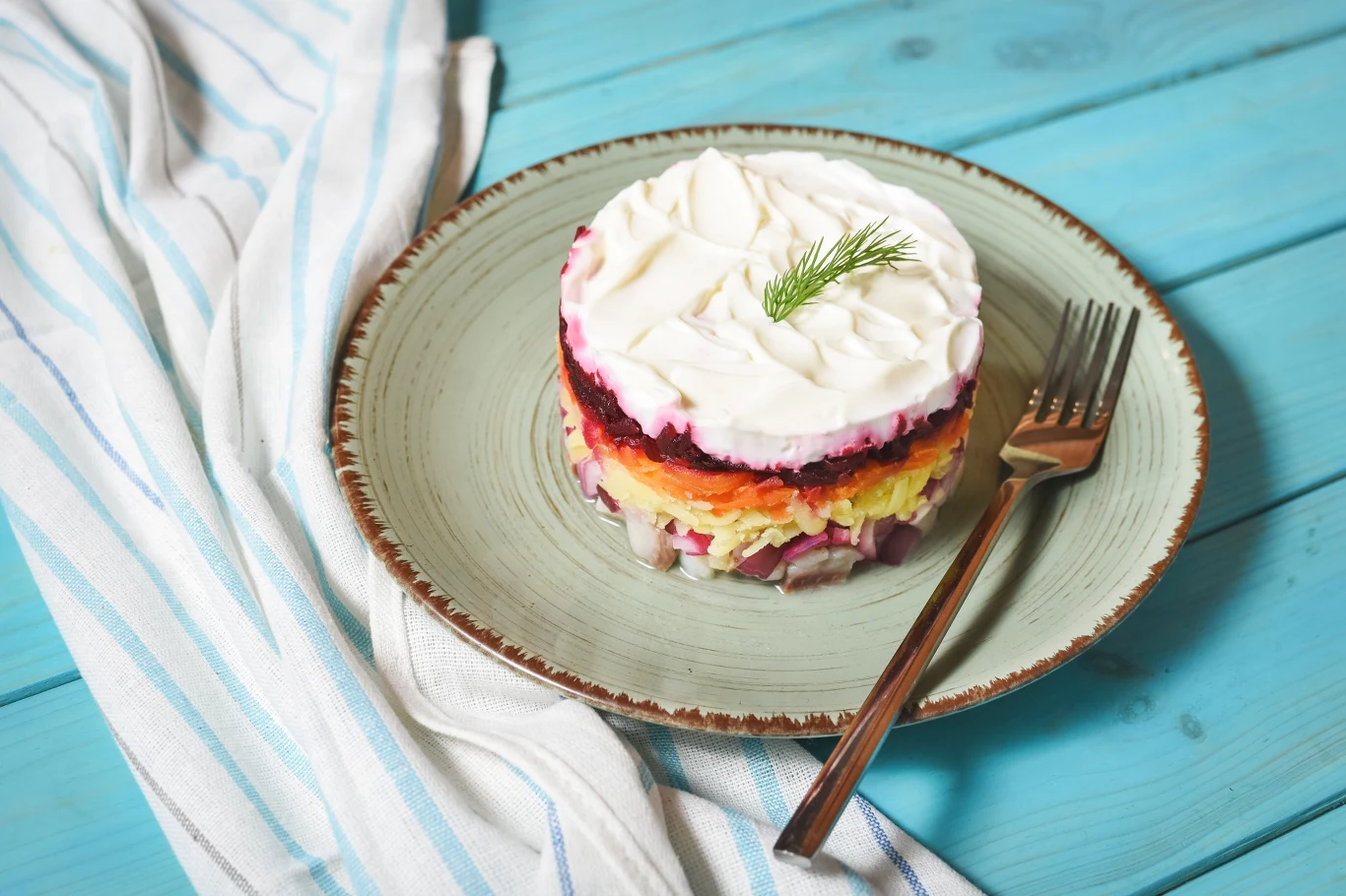 Ta sałatka śledziowa zrobi na wszystkich wrażenie. Doskonała propozycja na proszoną wigilię Warstwowa sałatka śledziowa z warzywami i majonezem, ułożona w okrągłej formie na talerzu, udekorowana koperkiem, obok leży widelec, a całość znajduje się na niebieskim drewnianym stole z białą ścierką w paski.