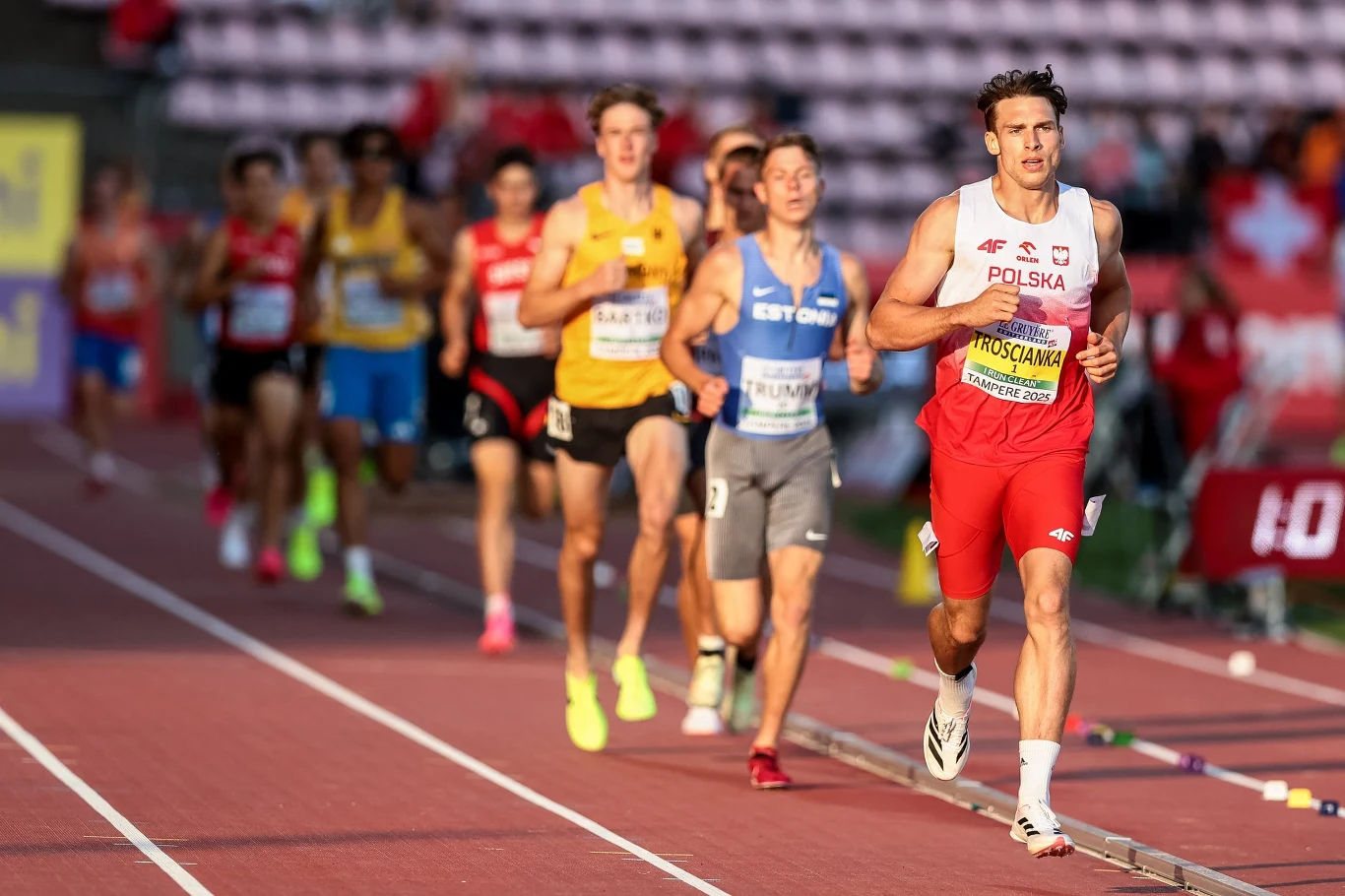 Hubert Trościanka w drodze po rekord świata. Tampere 2025 Grupa lekkoatletów biegnących po tartanowej bieżni stadionu, na czele zawodnik z Polski w biało-czerwonym stroju, pozostali uczestnicy w różnych barwach reprezentujących inne kraje, w tle trybuny oraz tablice zawodów.