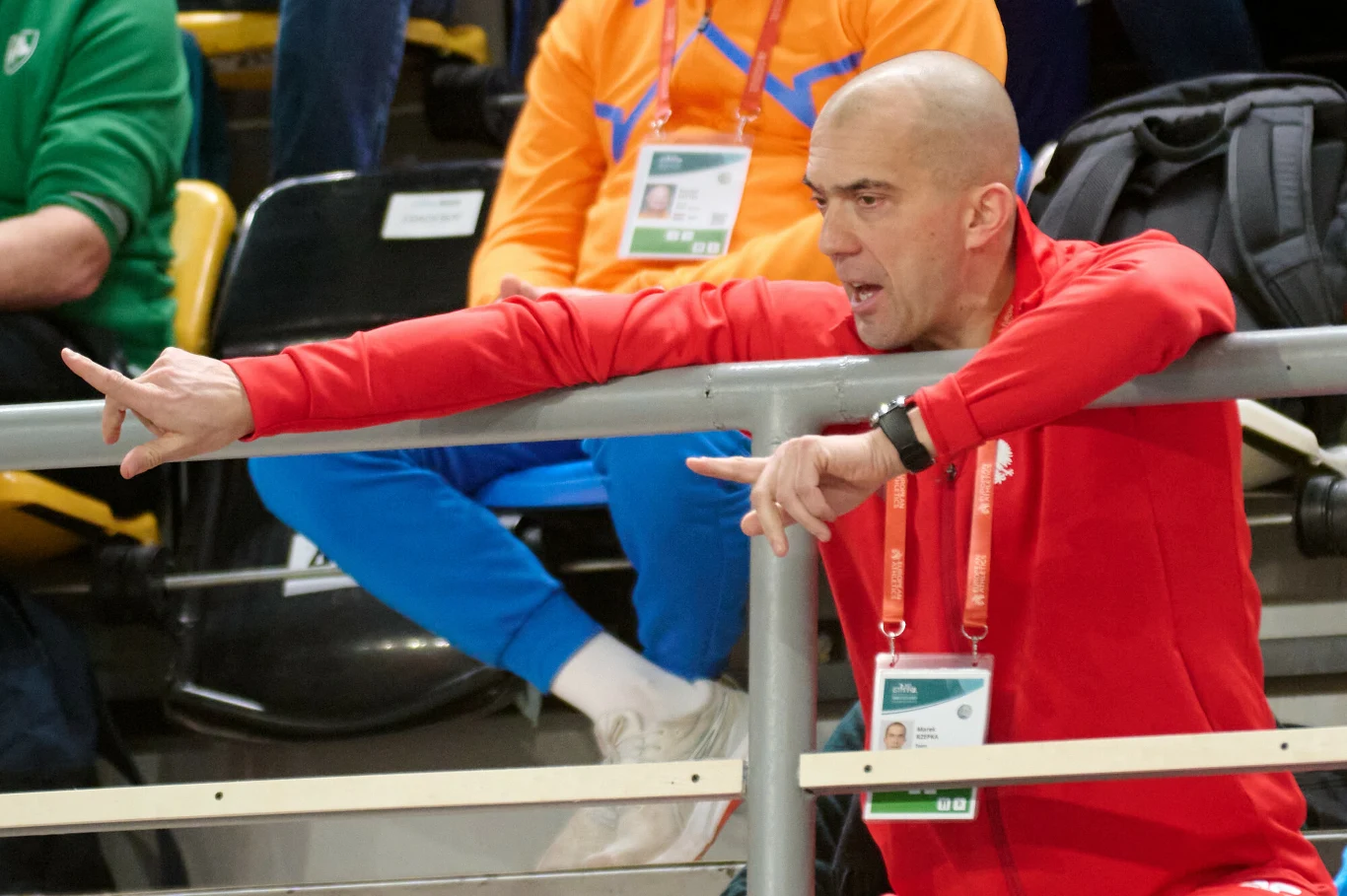 Trener Marek Rzepka Łysy mężczyzna w czerwonej bluzie energicznie gestykuluje, wskazując palcem poza kadr, siedząc na trybunach hali sportowej. W tle widoczna druga osoba w pomarańczowej bluzie i niebieskich spodniach.