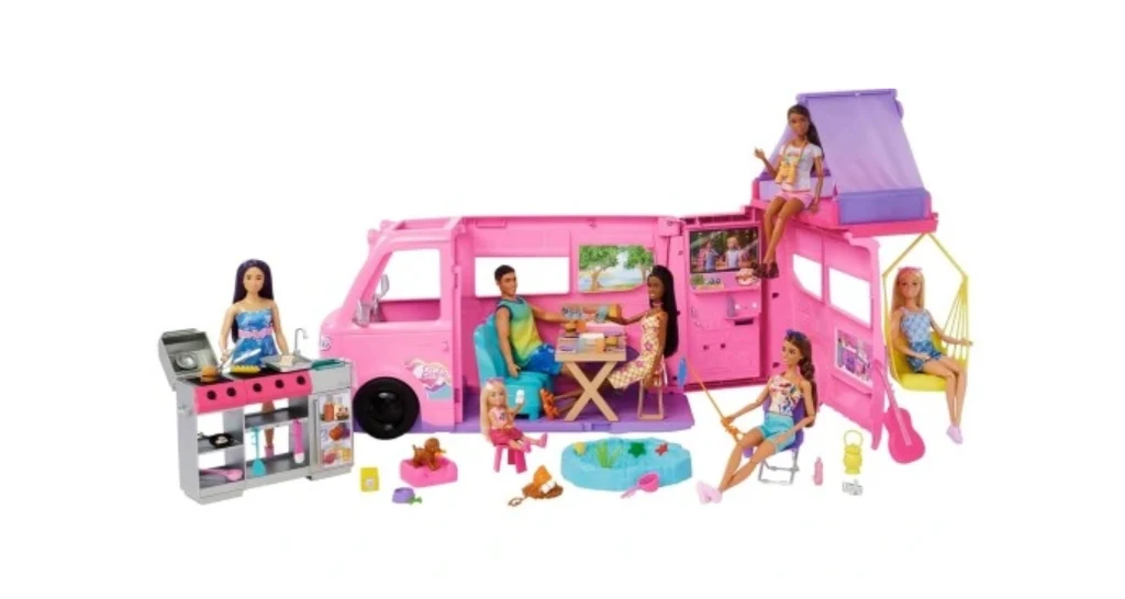 W zestawie Dreamcamper znajdziemy m.in. namiot na dachu, grill, jadalnię i wiszący fotel Różowy kamper zabawkowy rozłożony na części, wokół którego siedzi i bawi się kilka lalek. Na pierwszym planie widać zestaw akcesoriów do gotowania i basen dla lalek, a w środku pojazdu stół z krzesłami oraz aranżację przypominającą rodzinne wakacje.