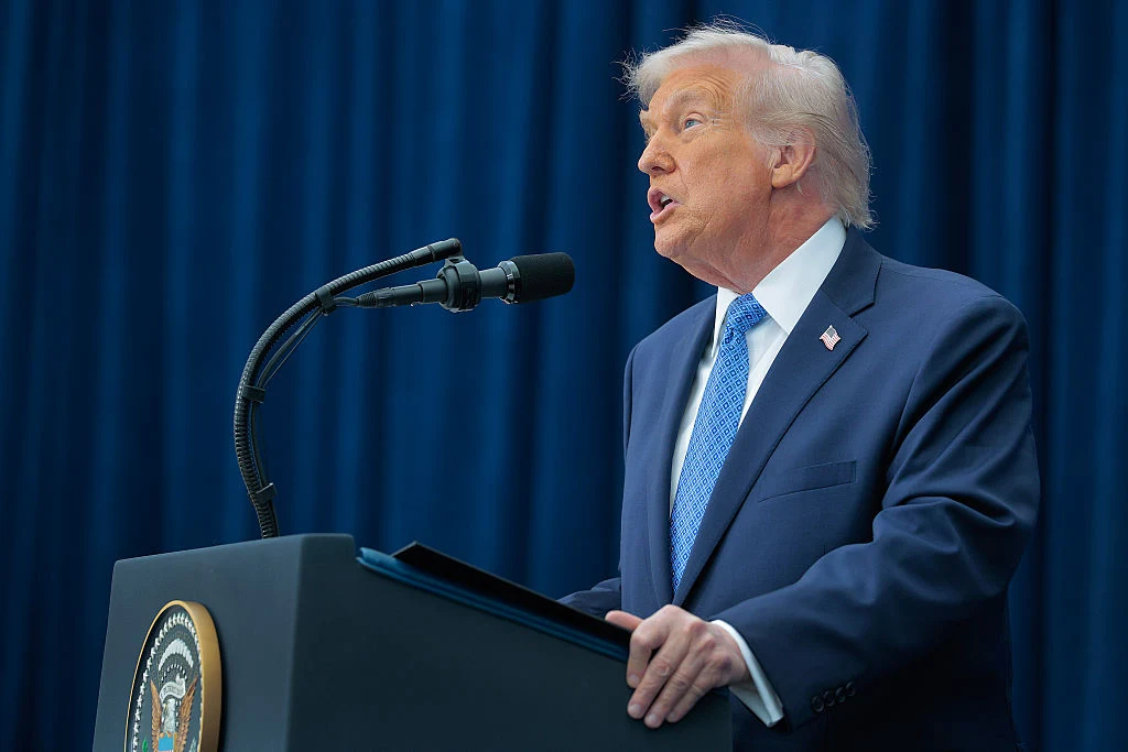 Prezydent USA Donald Trump Mężczyzna w garniturze mówi do mikrofonu stojąc przy mównicy z amerykańskim herbem prezydenckim, w tle ciemnoniebieska kurtyna.