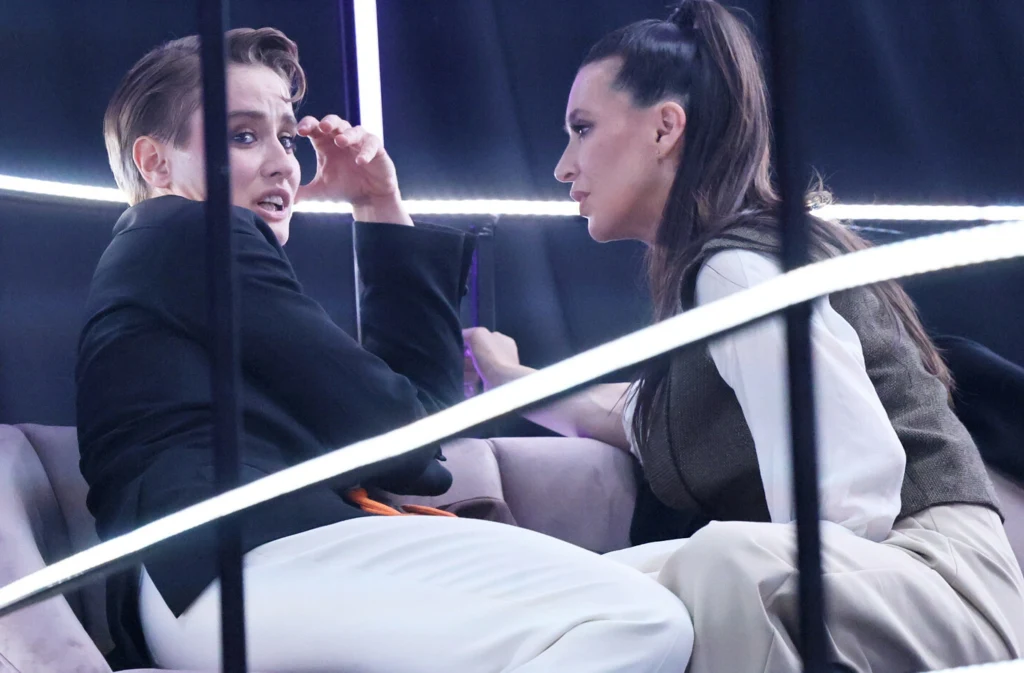 Dwie kobiety siedzące naprzeciw siebie na sofie w jasnych ubraniach, jedna z nich wydaje się zaskoczona, druga słucha z uwagą, całość oświetlona nowoczesnymi liniami świetlnymi.