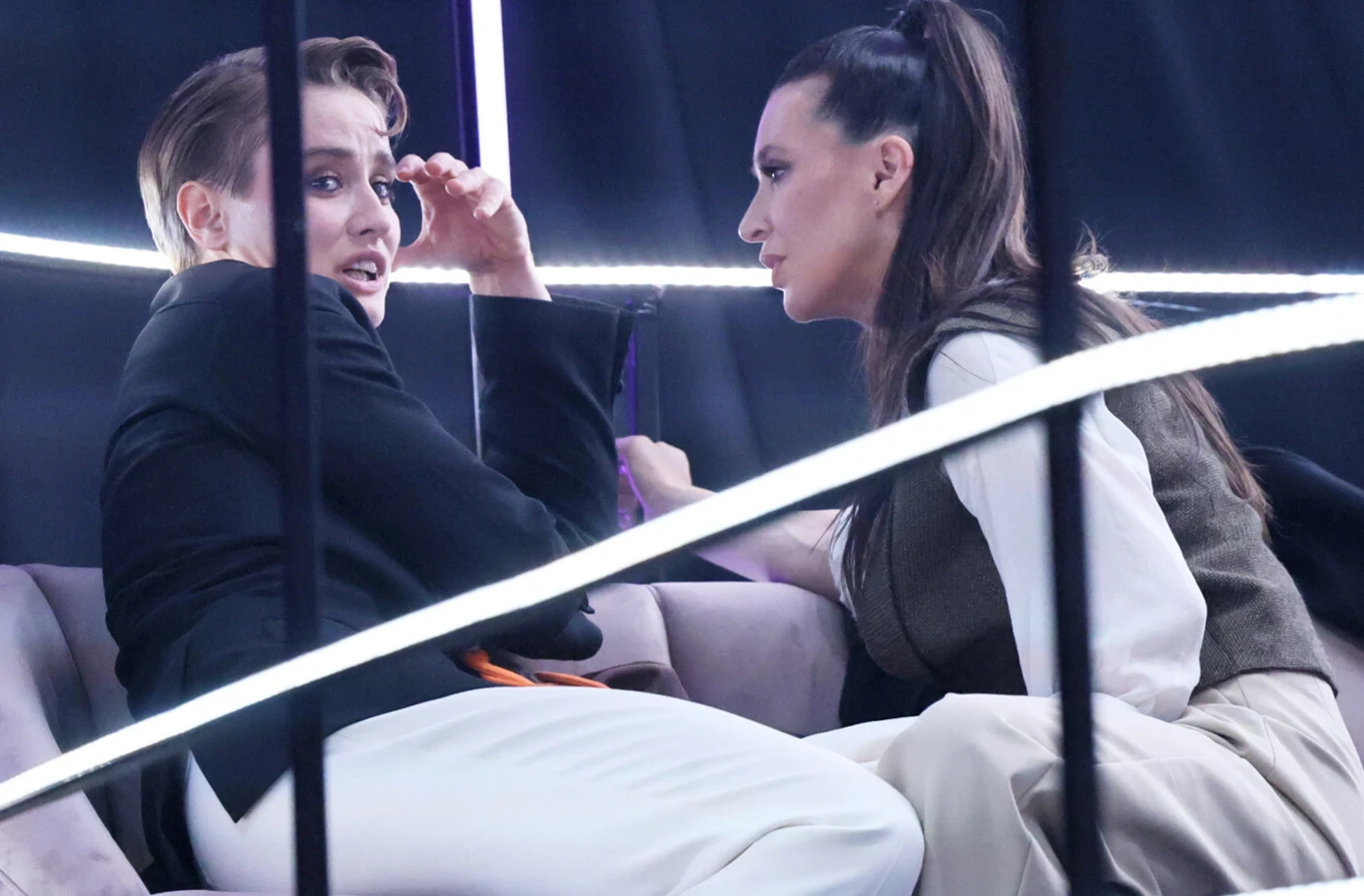 Katarzyna Zillmann, Janja Lesar Dwie kobiety siedzące naprzeciw siebie na sofie w jasnych ubraniach, jedna z nich wydaje się zaskoczona, druga słucha z uwagą, całość oświetlona nowoczesnymi liniami świetlnymi.