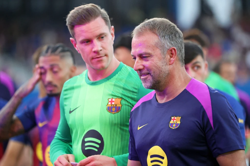 Hansi Flick i Marc-Andre ter Stegen Dwóch mężczyzn ubranych w stroje drużyny FC Barcelona, jeden w koszulce bramkarskiej, drugi w koszulce sztabu szkoleniowego, stoją obok siebie na tle innych zawodników przygotowujących się do meczu piłkarskiego.