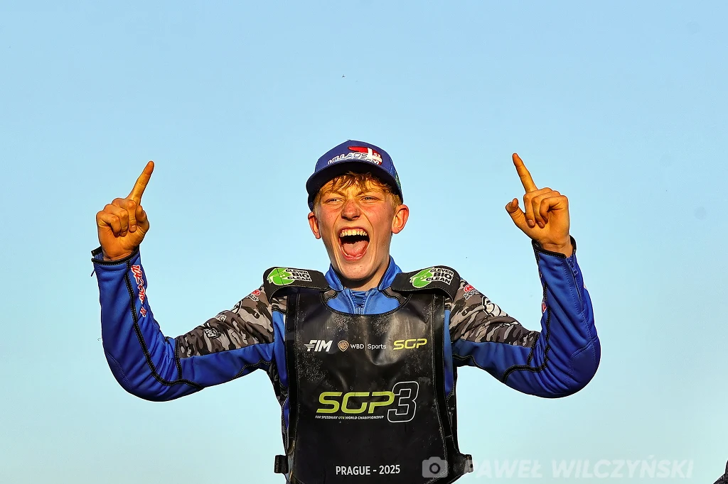 Młody zawodnik ubrany w sportowy strój żużlowy cieszy się z sukcesu, unosząc obie ręce z wystawionymi palcami wskazującymi, na tle jasnego nieba.