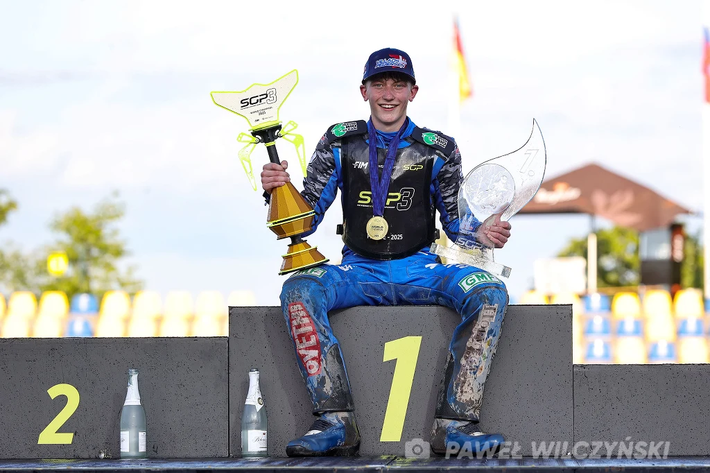 Młody zawodnik z szerokim uśmiechem siedzi na podium na pierwszym miejscu, trzyma w rękach dwa trofea, ubrany w strój wyścigowy pokryty błotem, w tle kolorowe elementy toru oraz rozmyte flagi.