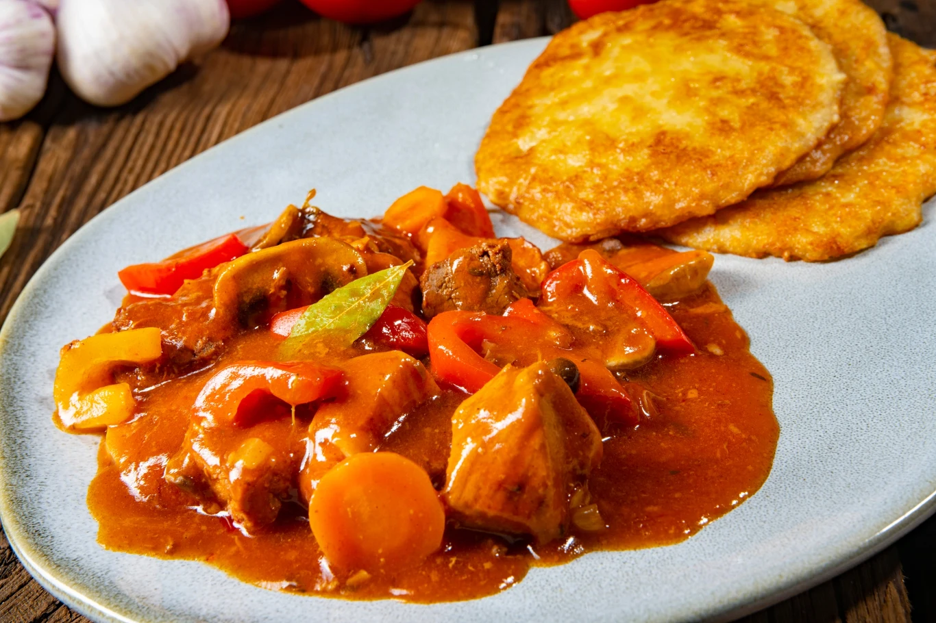 Placki ziemniaczane z gulaszem to idealne danie na zimowe chłody Placki ziemniaczane podane z gulaszem mięsnym, w którym widoczne są kawałki mięsa, marchew, papryka oraz gęsty, aromatyczny sos, całość ułożona na szarym talerzu na drewnianym stole, w tle widoczne główki czosnku i pomidory.