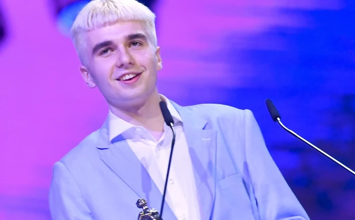 Młody mężczyzna w jasnoniebieskim garniturze, z jasnymi blond włosami, stoi za mównicą, trzyma statuetkę i uśmiecha się. W tle rozmyte, kolorowe światła sceniczne.