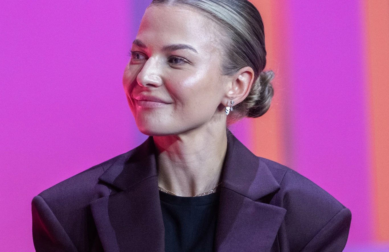 Anna Lewandowska Kobieta z upiętymi włosami ubrana w fioletową marynarkę i czarną bluzkę, delikatny makijaż oraz niewielkie kolczyki, jednolite różowo-fioletowe tło.