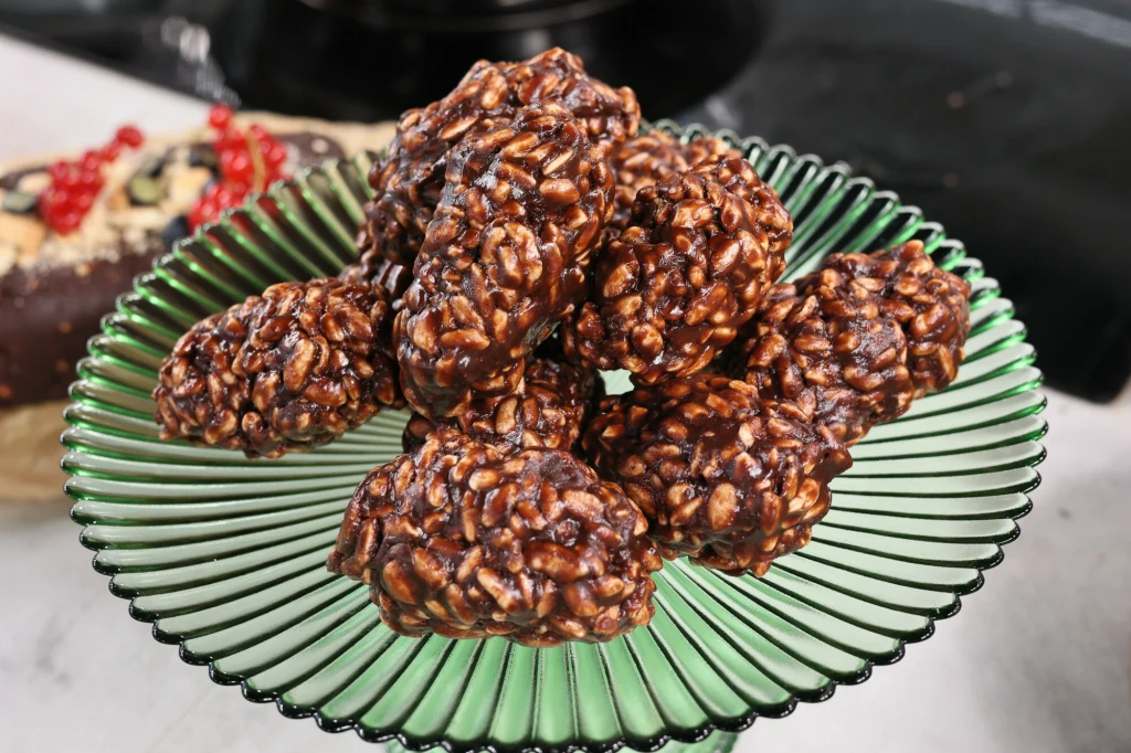 Stos uformowanych ciastek zbożowych w czekoladzie ułożony na zielonym szklanym talerzu, w tle widać fragment innego deseru.