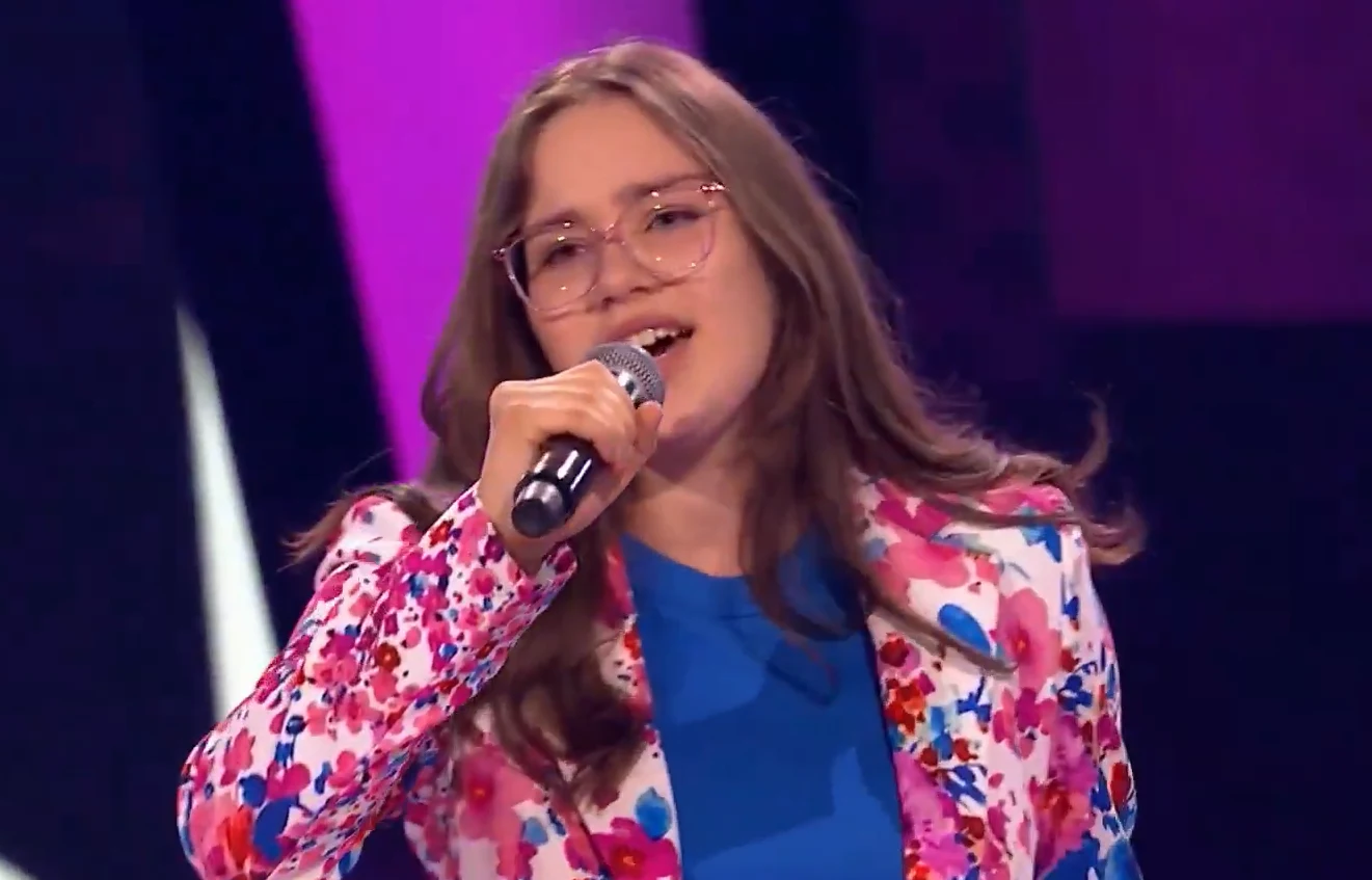 Ania Jarząbek w "The Voice Kids" w 2024 r. Młoda kobieta w okularach, ubrana w kolorową marynarkę z wzorem kwiatowym i niebieską bluzkę, śpiewa do mikrofonu na scenie oświetlonej różowym światłem.