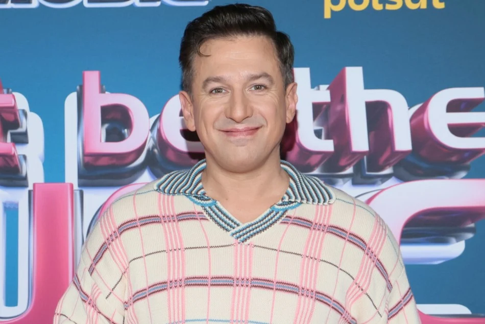 Maciej Rock zdradził, jak wyglądają jego święta Uśmiechnięty mężczyzna o ciemnych włosach stoi na tle tablicy z kolorowym napisem i logotypem, ubrany w jasny sweter w kratę z niebieskim kołnierzem.