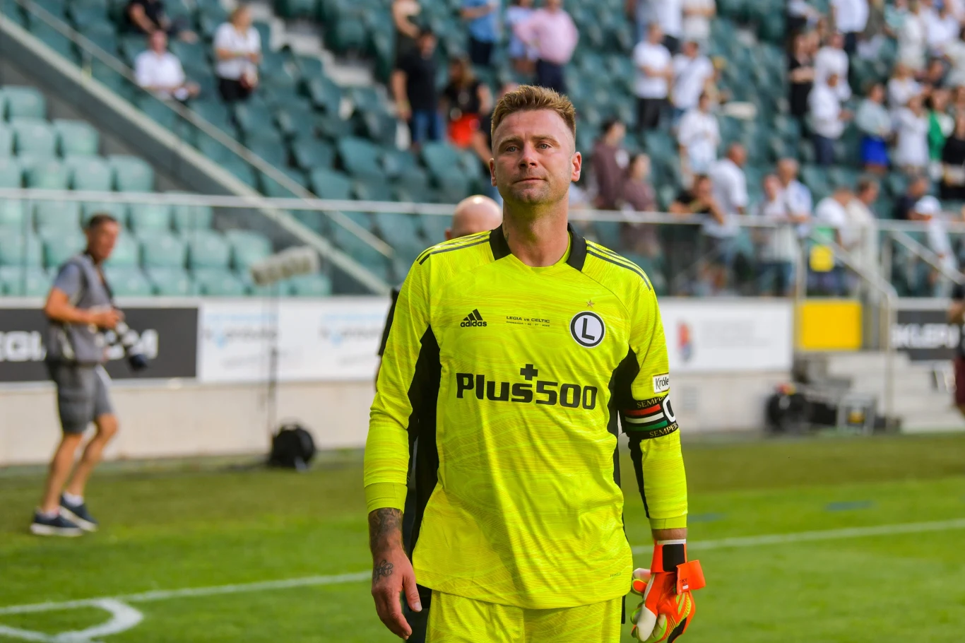 Artur Boruc Piłkarz w żółtym stroju bramkarskim na stadionie, z opaską kapitana na ramieniu, trzymający rękawice w jednej dłoni, w tle widać trybuny oraz rozproszonych kibiców.
