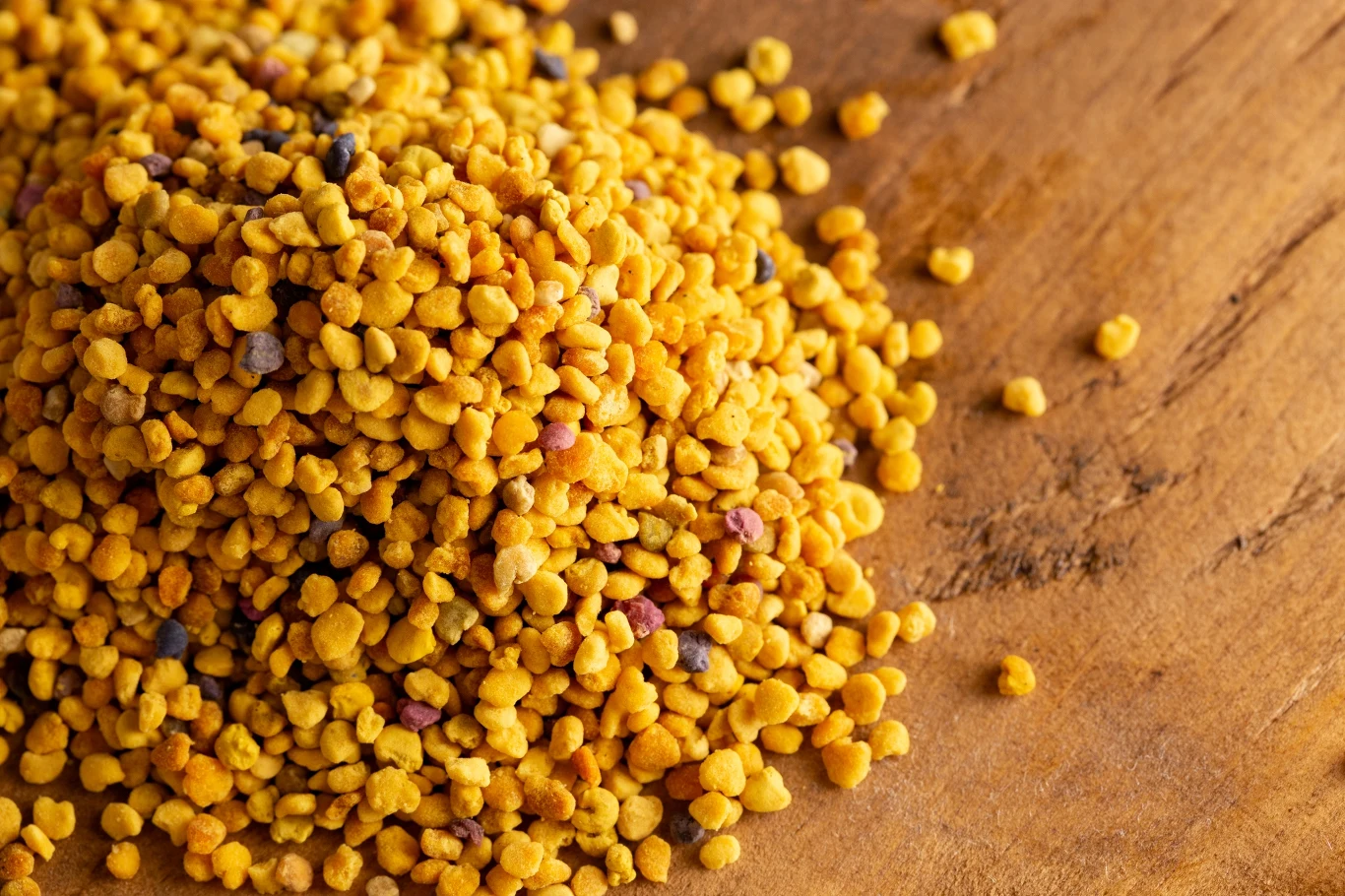 GIS wydał ostrzeżenie publiczne dotyczące żywności: wysoki poziom alkaloidów pirolizydynowych w określonej partii pyłku kwiatowego Stos pyłku pszczelego o żółtej i pomarańczowej barwie rozsypany na drewnianej powierzchni, widoczne drobne i nieregularne granulki.