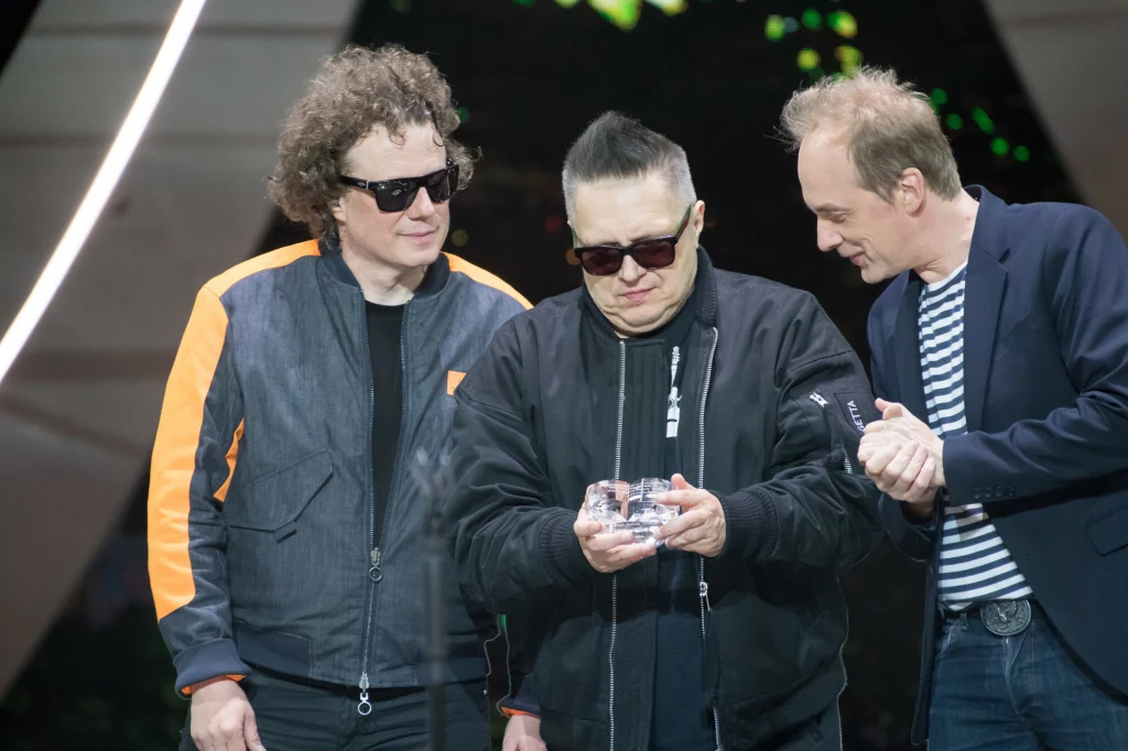 Muzycy T.Love w 2022 r. - od lewej Sidney Polak, Muniek Staszczyk i Jan Benedek. Perkusista i gitarzysta są już poza zespołem Trzech mężczyzn w ciemnych okularach na scenie, jeden z nich trzyma szklaną nagrodę, pozostali spoglądają na nią z zaciekawieniem, tworząc atmosferę skupienia i zainteresowania.