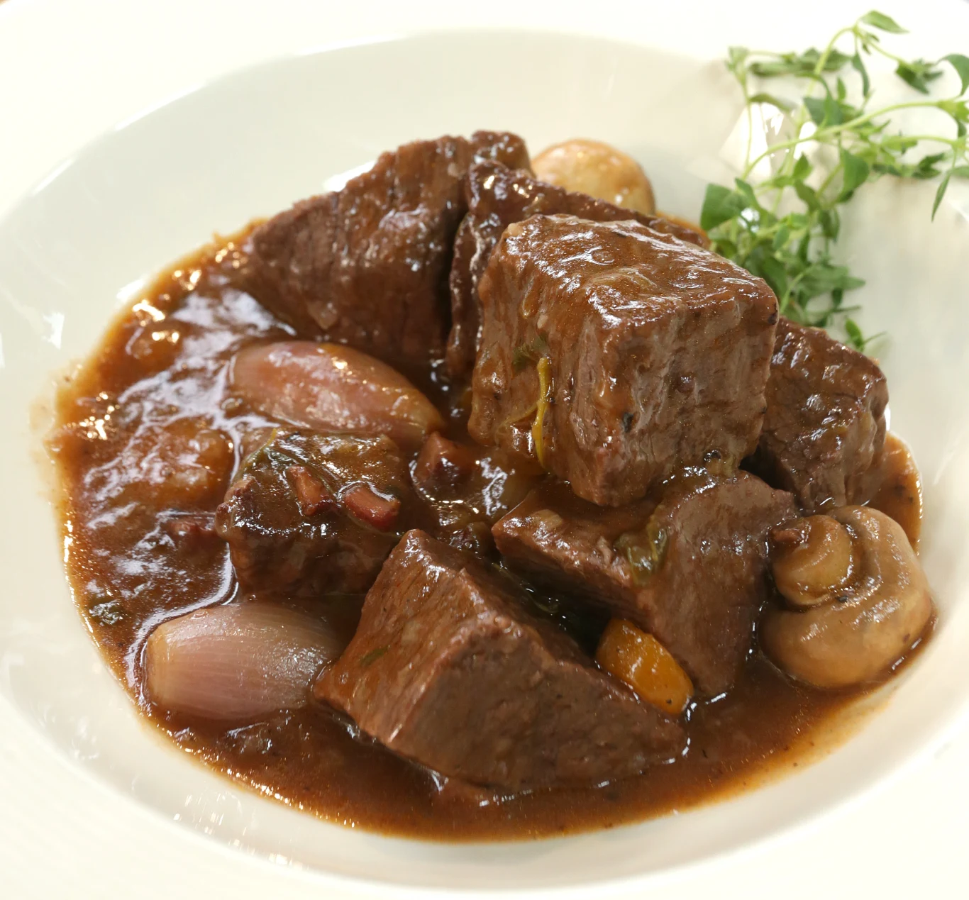 Duże kawałki duszonej wołowiny zanurzone w gęstym, ciemnym sosie, z dodatkiem pieczarek i całych małych cebulek, udekorowane świeżymi ziołami.