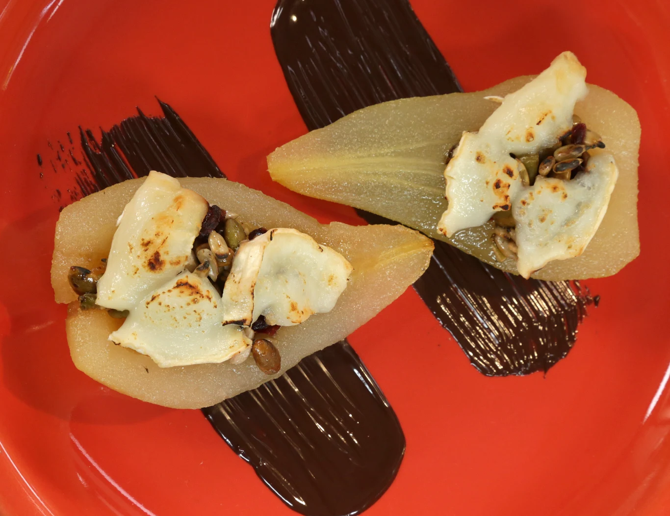 Dwie połówki gruszki, zapieczone z serem oraz pestkami, ułożone na czerwonym talerzu z dekoracyjnymi pociągnięciami ciemnej czekolady.