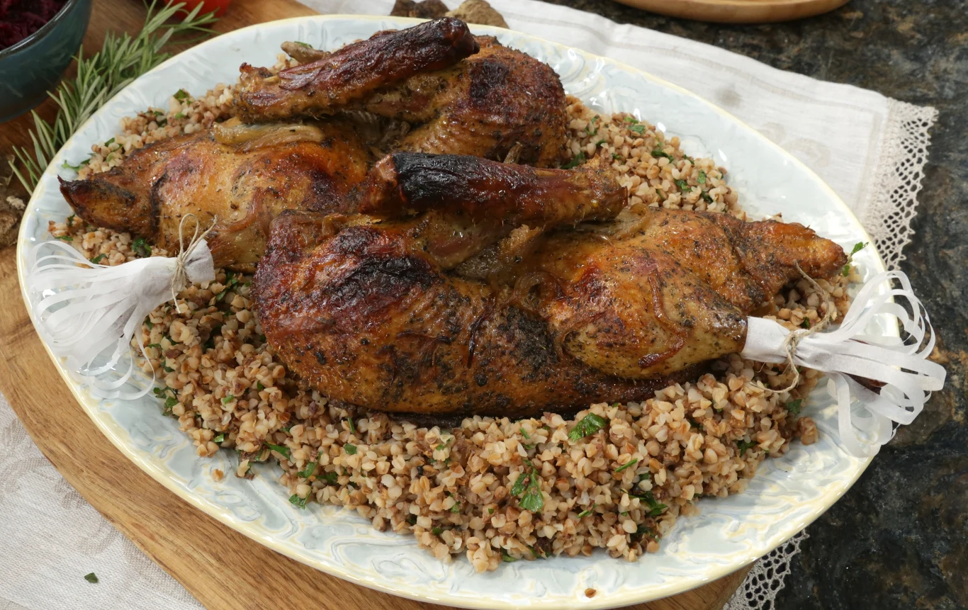 Pieczony kurczak serwowany na dużym półmisku na podkładzie z kaszy gryczanej, ozdobiony ziołami oraz dekoracyjnymi papierowymi mankietami na końcach nóg.