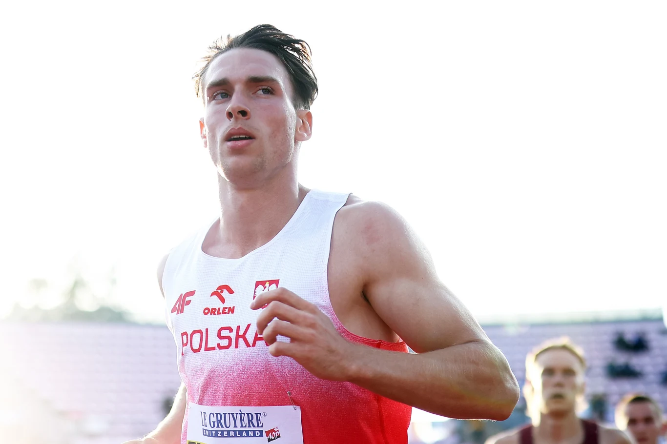 Hubert Trościanka Młody sportowiec w stroju reprezentacji Polski biegnie na stadionie lekkoatletycznym, wokół widoczni są inni zawodnicy i kibice w tle.