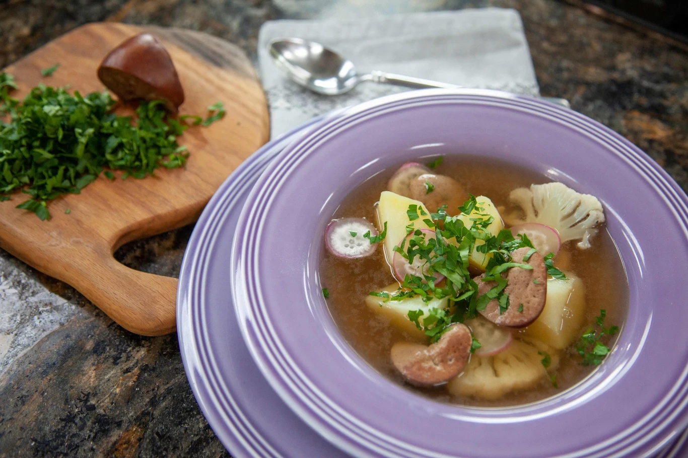 Talerz z zupą zawierającą plasterki ziemniaka, kalafior, kawałki podrobów oraz świeżą natkę pietruszki, obok na desce znajduje się posiekana natka pietruszki i połowa podrobów, w tle leży łyżka na serwetce.